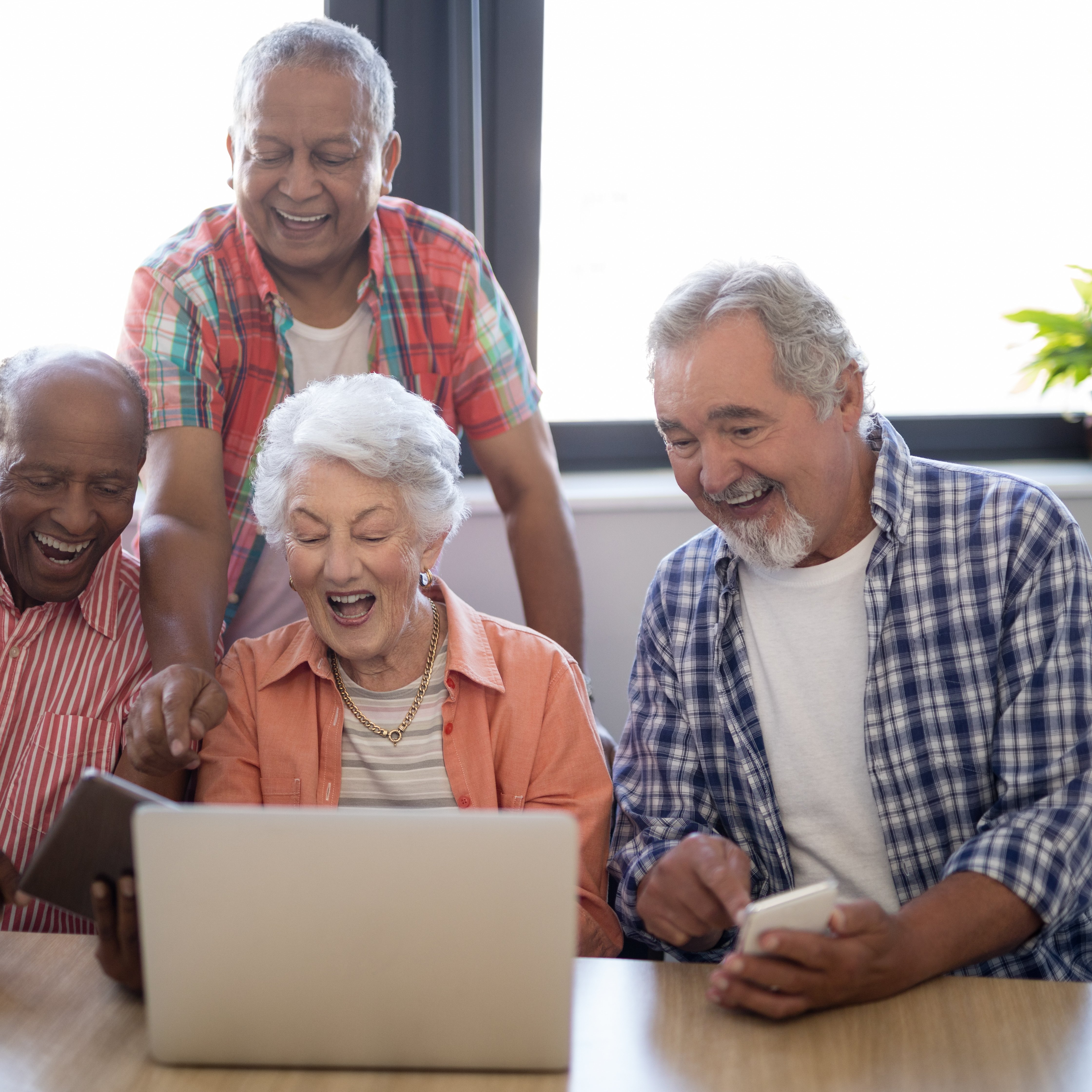 Happy senior people using technology while sitting at table against window in nursing home